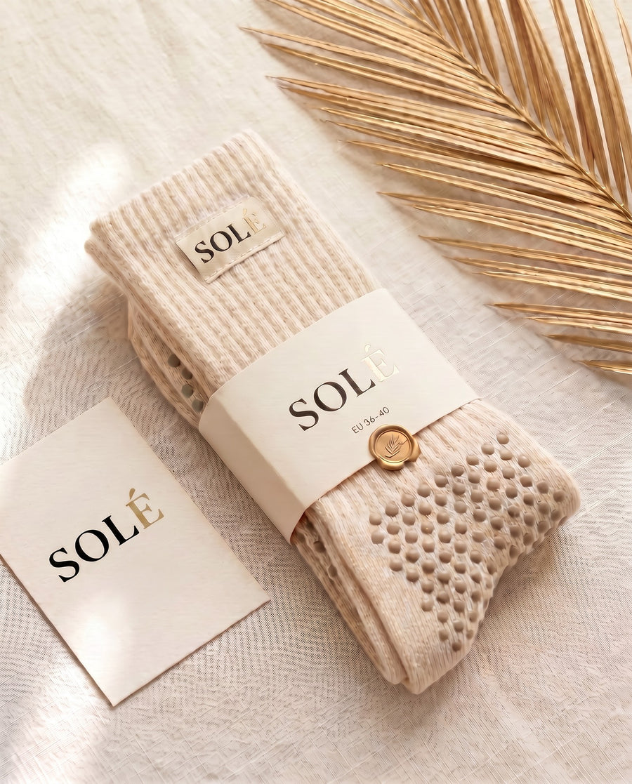 A pair of SOLÉ cream grip socks with tonal taupe grip dots, wrapped in a SOLÉ belly band with a QR care card